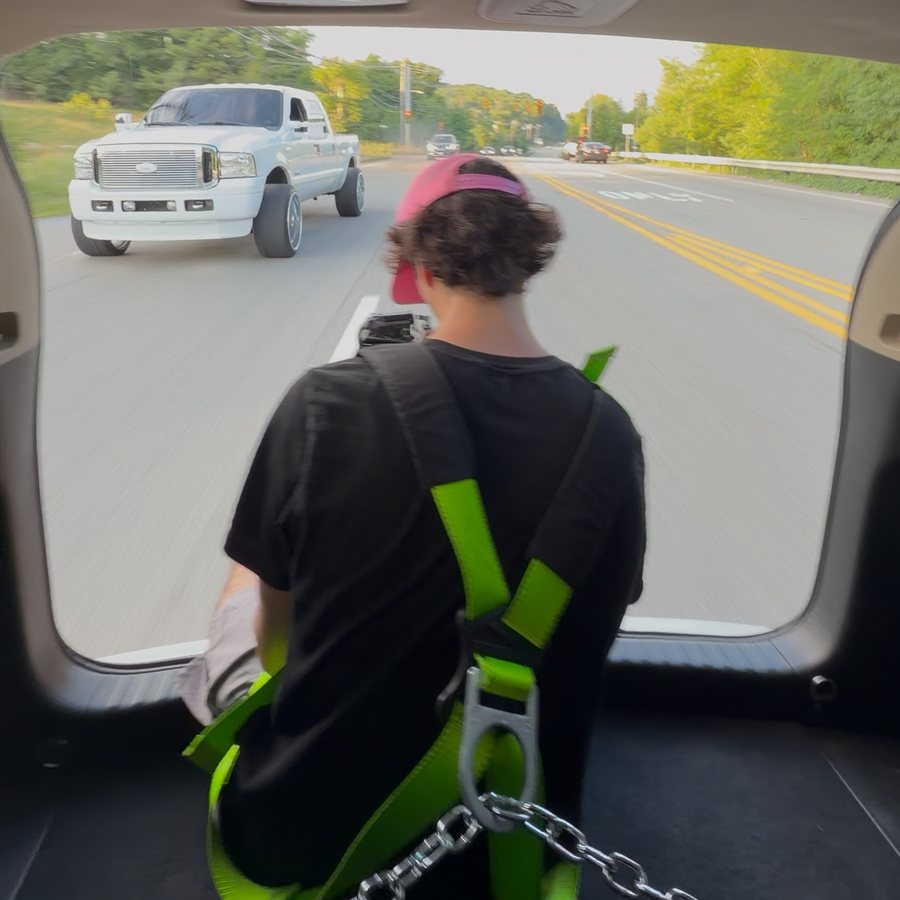 Gianni harnessed in the back of a moving car, shooting a white Ford truck on the road